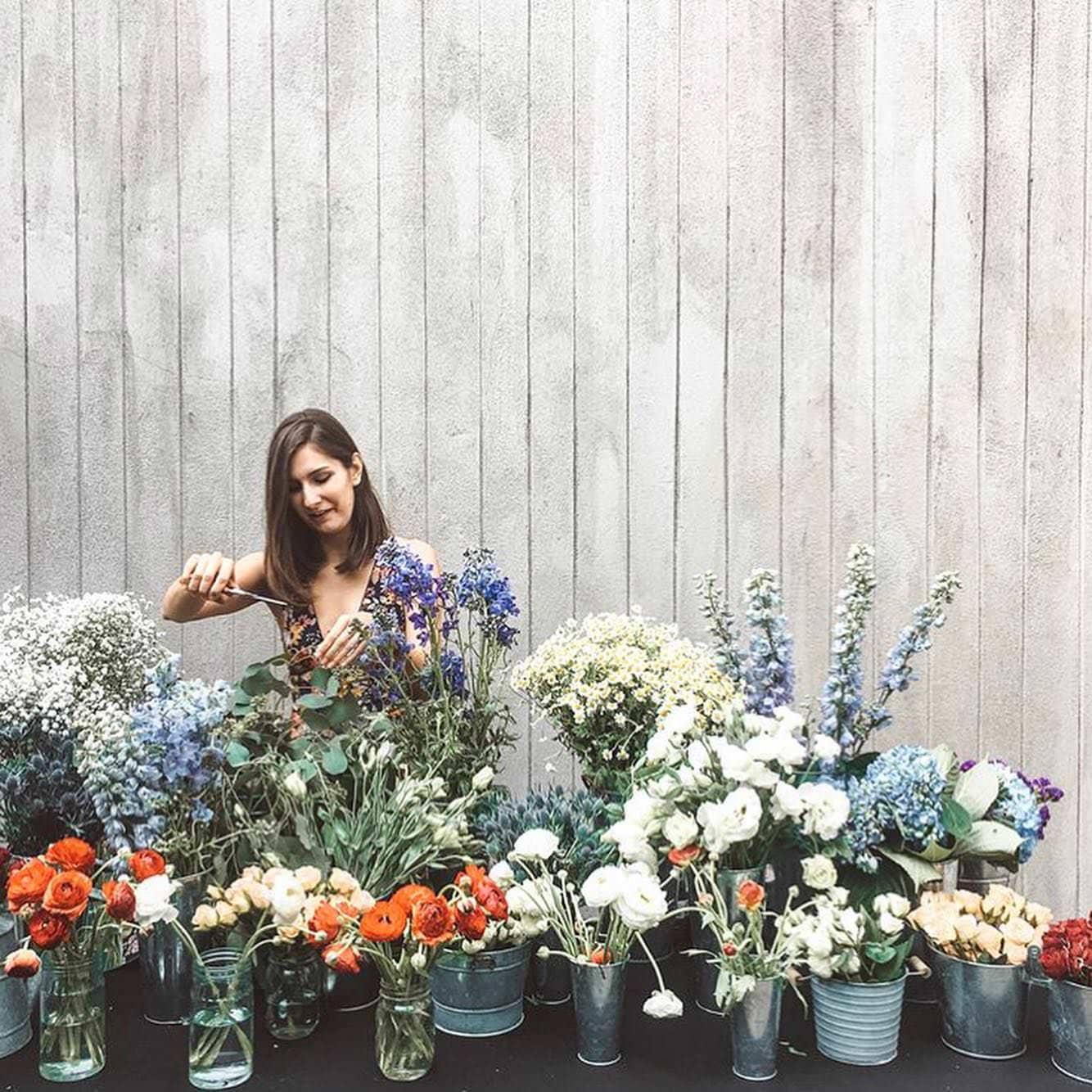 A woman arranges flowers in vases and metal buckets on a table in front of a light gray wooden wall, turning her workplace into a burst of color with red poppies, white roses, blue delphiniums, and baby's breath. She appears focused on her business. - Flower Gypsies - Luxury Floral Design, Los Angeles CA