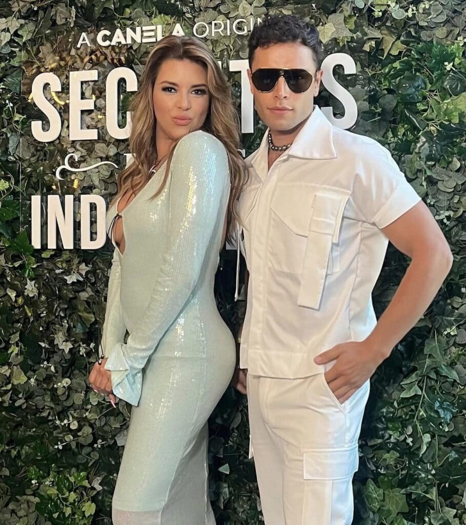 Two people posing at event with foliage background.