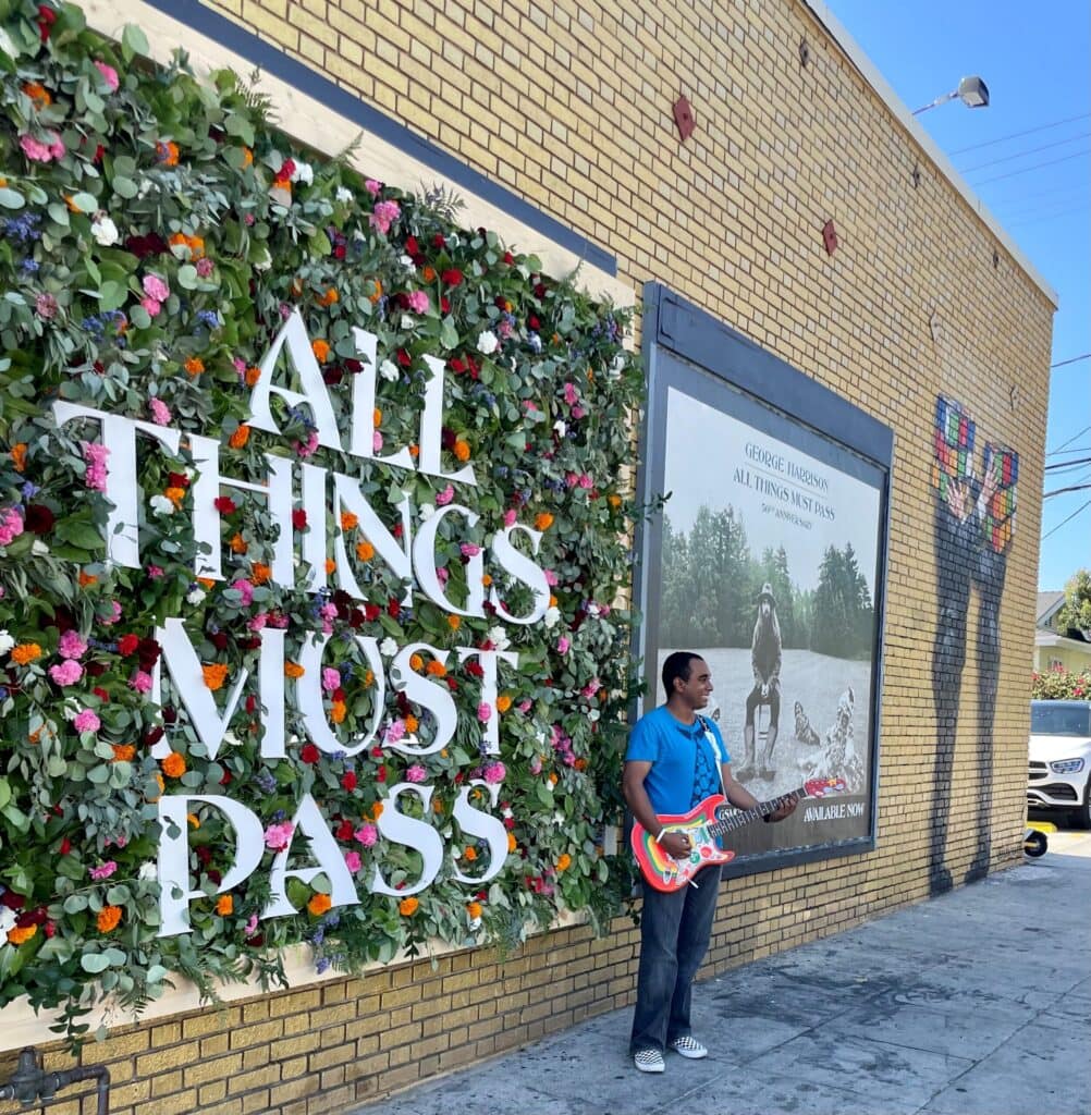 Man with guitar by floral 'All Things Must Pass' wall.